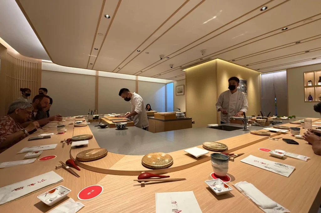 The wide, minimalist interior of a Japanese restaurant features a large, U-shaped light wood counter where several patrons are seated and interacting with chefs wearing white uniforms and black masks. The elegant space is illuminated by soft, recessed lighting, and the table is meticulously set with individual plates, cups, and napkins, reflecting a calm, sophisticated dining experience.