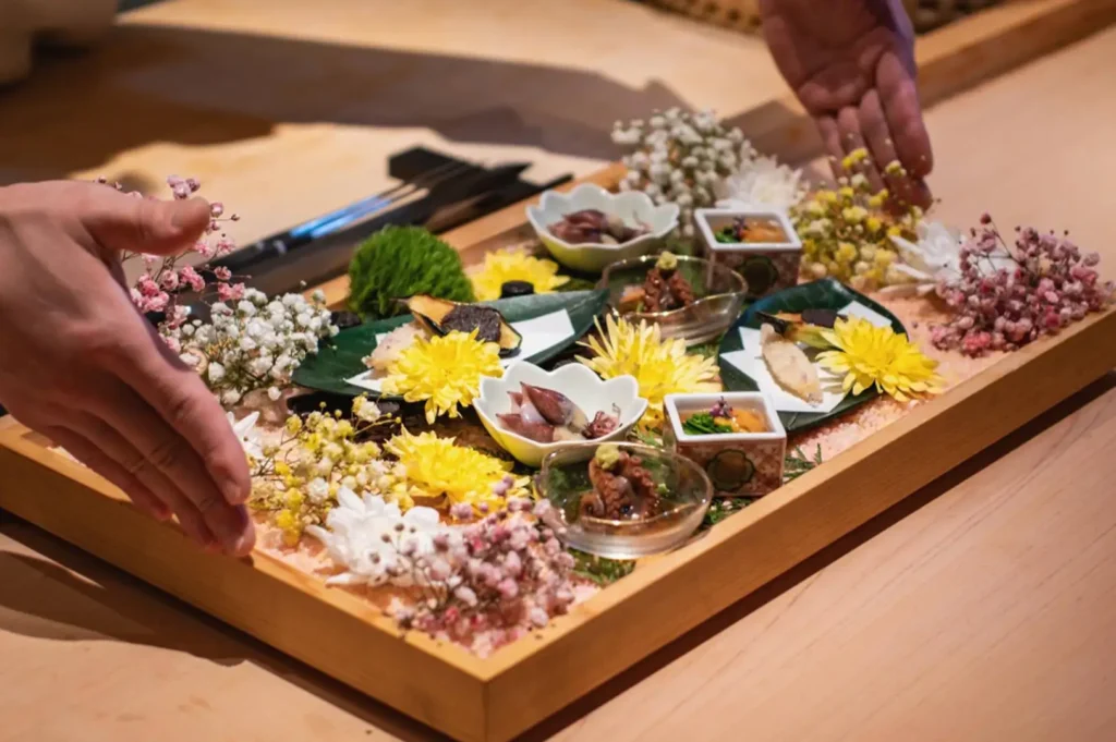 A chef’s hands present a large, rectangular wooden tray bedded with pink Himalayan salt and filled with an elaborate omakase appetizer arrangement, featuring small ceramic bowls of firefly squid, sea urchin, braised octopus, grilled fish, and delicate tempura, all artfully decorated with vibrant yellow chrysanthemums and colorful sprigs of dried baby’s breath flowers.