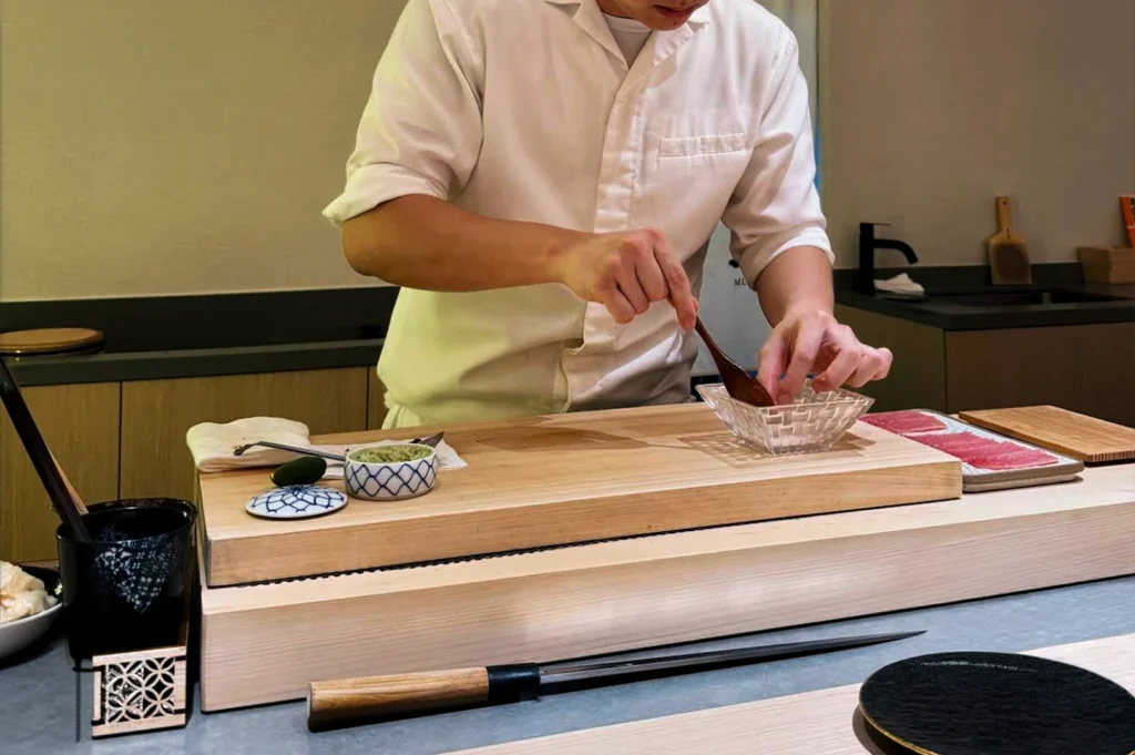 A chef wearing a crisp white coat uses a small wooden spoon to work with an ingredient in a glass bowl, standing behind a large wooden prep counter equipped with a long knife and various small ceramic vessels for seasoning.