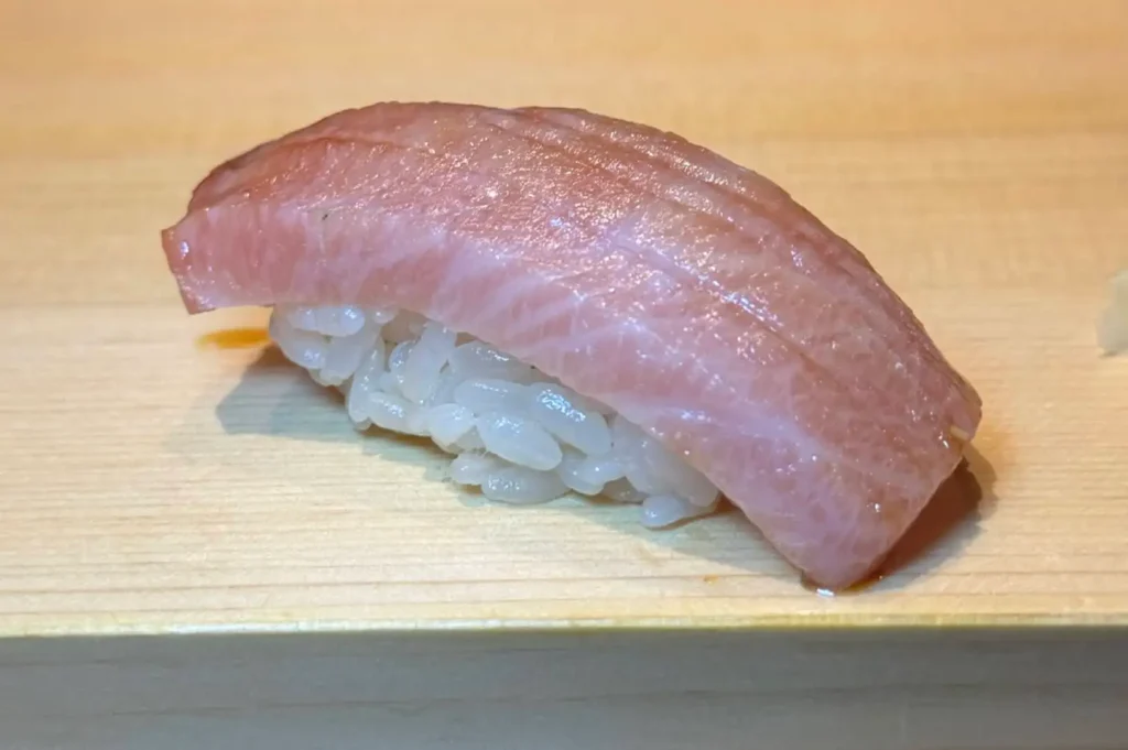 A detailed macro shot highlights a single piece of premium nigiri sushi resting on a smooth, light-colored wooden counter block. The sushi features a perfectly shaped mound of white vinegared rice topped with a thick, curved slice of pale pink, fatty tuna sashimi, likely otoro or chutoro. The fish displays intricate white marbling and fine scoring marks along its glistening surface, with a faint hint of soy glaze at its base.