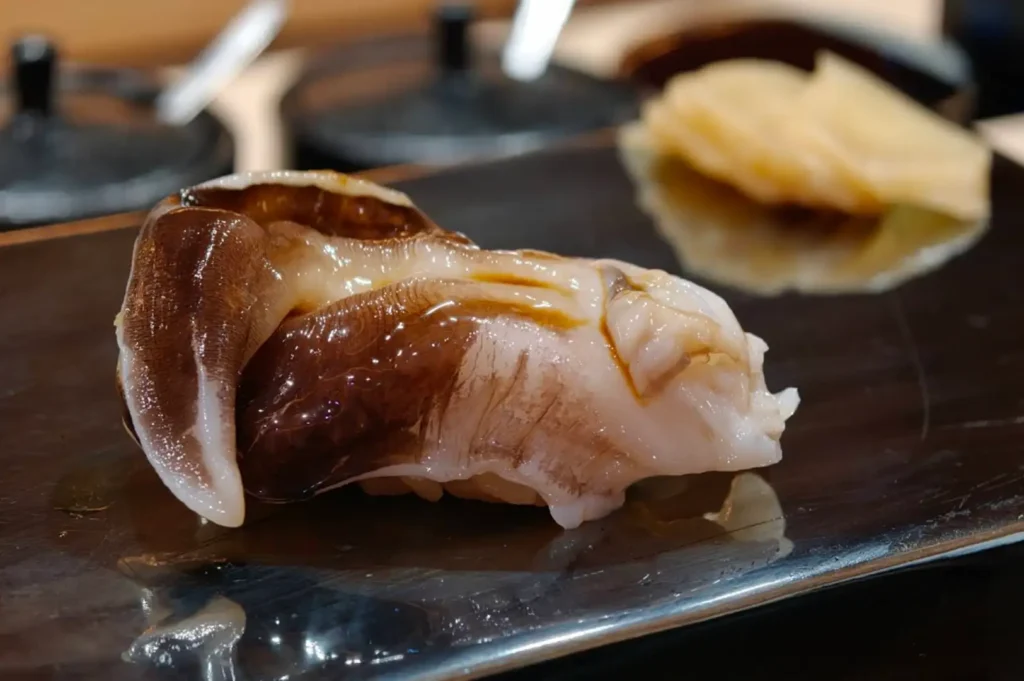 A detailed macro shot of a piece of fresh, glossy surf clam (torigai) nigiri sushi brushed with savory tare sauce, displayed on a dark rectangular serving platter next to thinly sliced pickled ginger.