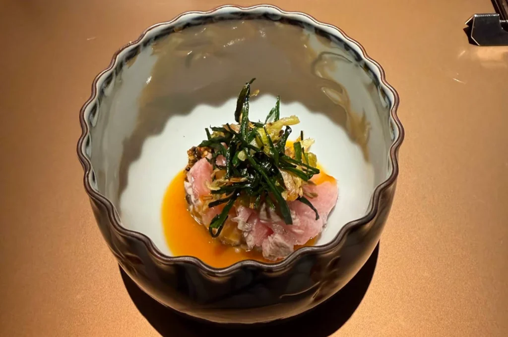 A top-down, close-up shot of an artistic Japanese dish served in a deep ceramic bowl with a wavy, dark-glazed rim and a smooth white interior. Thin, delicate slices of pale pink sashimi are neatly fanned out in a vibrant, bright orange egg yolk sauce at the bottom of the bowl. The fish is crowned with a high nest of finely shredded, crisped green scallions and herbs, catching the overhead spotlight on a bronze-toned counter.