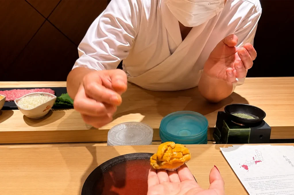 A sushi chef in a white uniform and face mask carefully places a piece of sea urchin (uni) nigiri into a customer's hand, while on the wooden counter nearby, there is a small bowl of rice, a menu with a map of Japan, and various small serving containers.