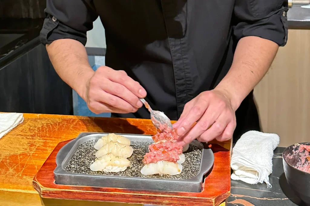An eye-level shot captures a chef from the chest down, dressed in a sleek black short-sleeved uniform, meticulously assembling sushi at a wooden counter. Using a small spoon held in his right hand, he carefully places a mount of finely chopped, pinkish-red tuna tartare infused with salmon roe onto a piece of sushi rice, while his left hand stabilizes the preparation. In front of him, a textured grey rectangular tray sits on a wooden base, holding two pieces of seared white scallop nigiri alongside the piece being prepared, flanked by a folded white towel on the side.