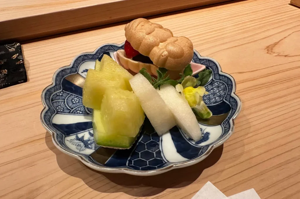 A colorful dessert served on a scalloped blue-and-white porcelain plate features sliced green melon, fresh white pear, a small edible yellow blossom, and a peanut-shaped monaka wafer filled with red bean paste and fresh fruit.