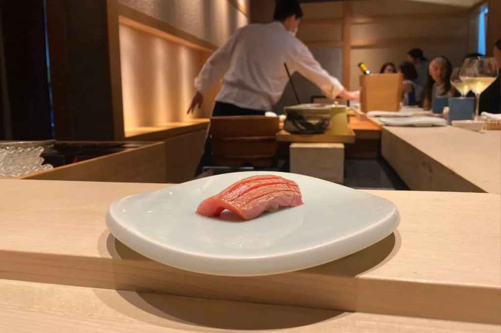 In the background of a refined, minimalist dining space, a chef is partially visible while working behind a long, light-wood counter. In the foreground, a single, perfectly prepared piece of tuna nigiri sits on a simple white plate, serving as the focal point of the image and capturing the serene and focused atmosphere of the omakase dining experience.