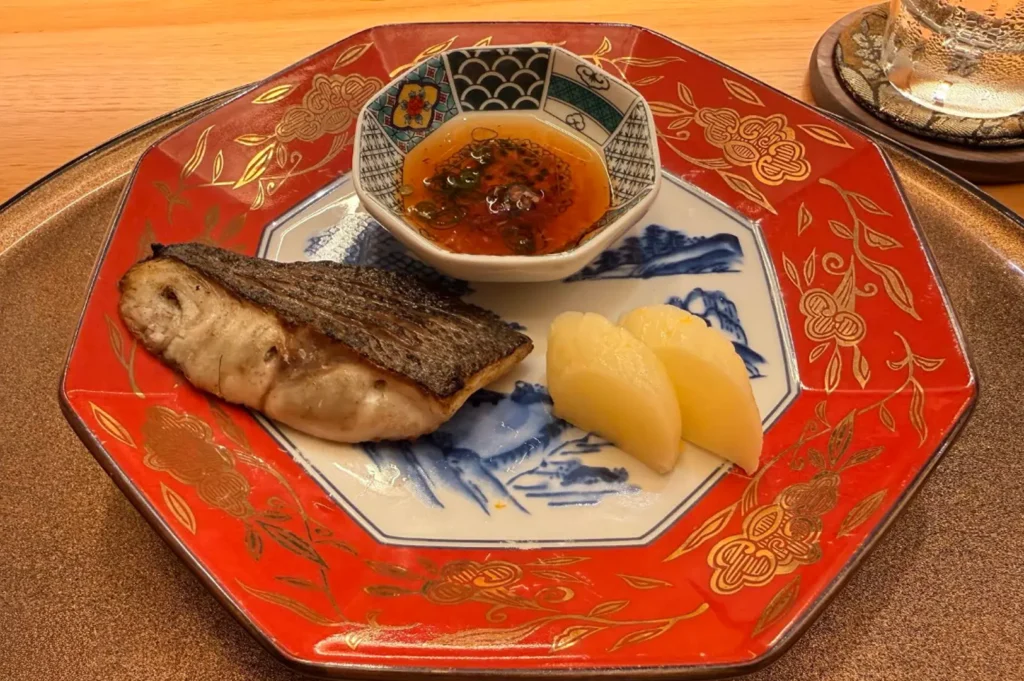 A piece of expertly cooked, flaky white fish is served on an ornate, hexagonal red and gold plate with intricate traditional patterns, accompanied by a small bowl of savory dipping sauce and two neatly cut wedges of pale, cooked root vegetable.