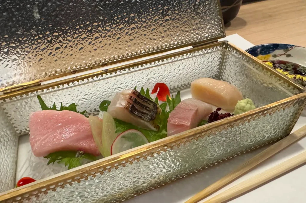 A selection of fresh sashimi artfully presented inside an open, rectangular vintage-style glass box with a hinged golden metal frame. The box sits on shredded daikon radish and holds five pieces of raw fish and seafood, including pale pink fatty tuna, white fish with seared skin, yellowtail, and a translucent scallop. The dish is garnished with green shiso leaves, thin radish slices, red edible flower petals, a dollop of fresh green wasabi, and a tiny mound of purple microgreens, with a pair of light wooden chopsticks resting alongside the box.