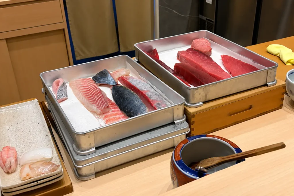 An omakase chef's prep station showcases two metal trays holding pristine cuts of fresh fish, including deep red tuna blocks and silver-skinned fillets. Next to the trays sits a ceramic pot with a wooden sauce brush, along with a plate displaying prepared sushi pieces like sweet shrimp and squid.