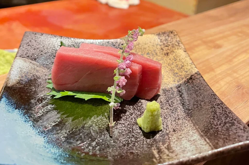 A close-up, high-angle shot of two thick, vibrant pieces of sashimi resting on a textured, dark ceramic plate. A delicate sprig of purple shiso flowers is placed gracefully across the fish, accompanied by a small dollop of bright green wasabi, set against a warm, wooden counter background.