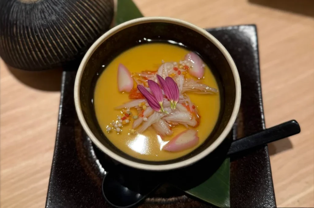 A small, elegant bowl of Japanese egg custard, or chawanmushi, is served in a dark, minimalist dish and topped with shredded crab, delicate purple flower petals, and tiny, colorful grains. The bowl is placed on a dark, square ceramic plate, which is set against a wooden table, and a black spoon rests beside it, showcasing a refined and artistic culinary presentation.