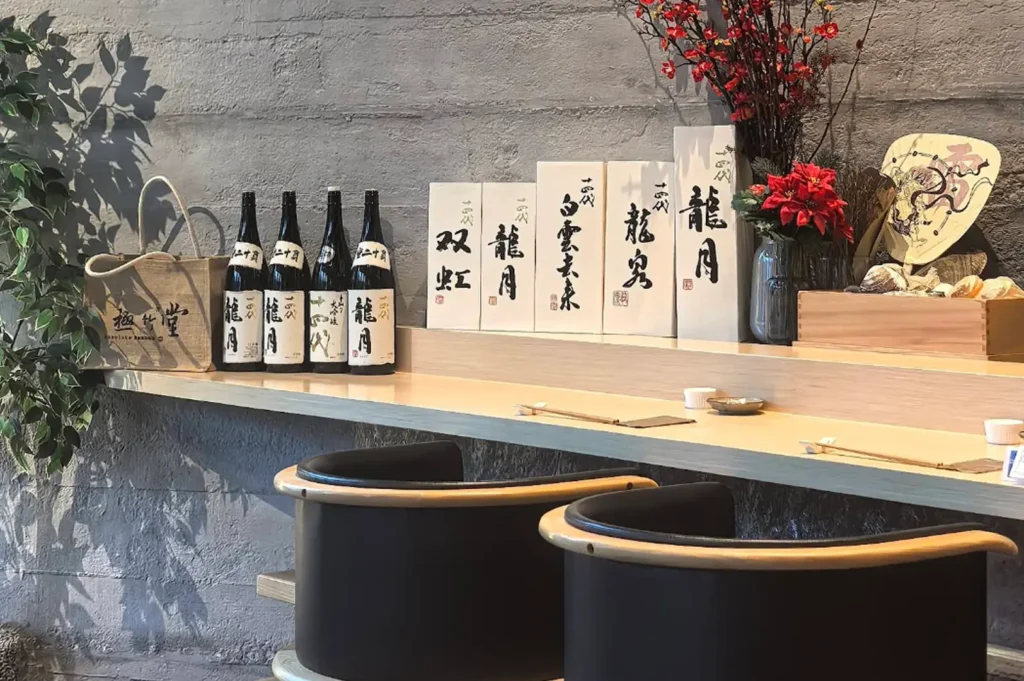 A wooden counter display features a collection of Japanese sake bottles arranged alongside tall, white wooden boxes inscribed with calligraphy, while a vase of red flowers and a decorative fan add to the refined aesthetic.