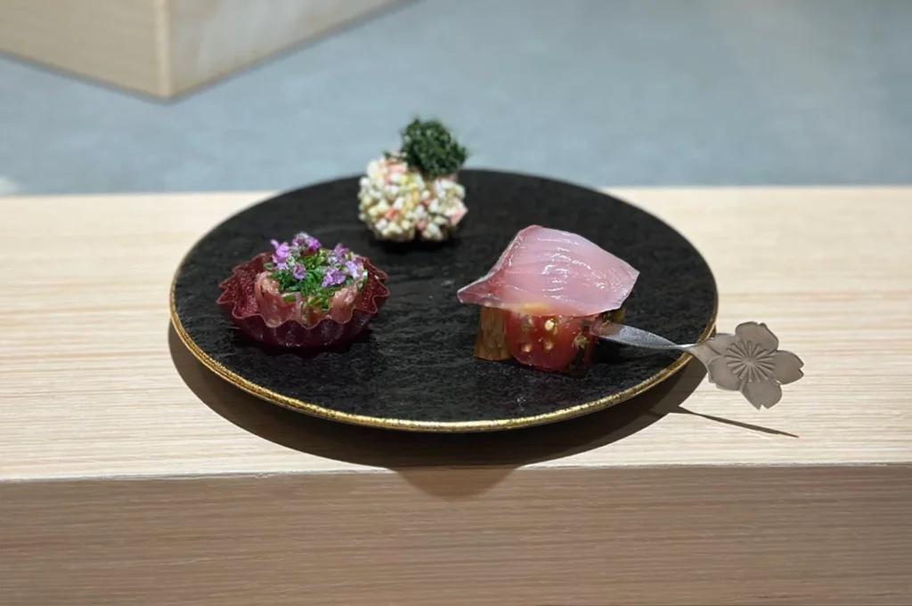 A black textured plate holds two small, intricate appetizers, including a tartlet topped with herbs and a piece of raw fish resting on a slice of tomato, served with a decorative metal utensil shaped like a flower petal.