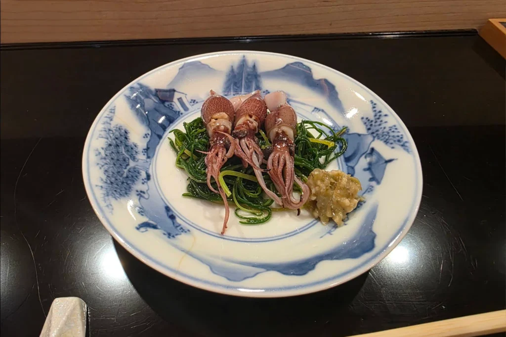 A top-down shot showcases an elegant culinary dish served on a traditional white ceramic plate with intricate blue landscape illustrations. Three small, whole cooked firefly squids (hotaru ika) with glossy, purple-brown bodies are neatly arranged side-by-side atop a bed of vibrant green sea chives. A small mound of textured, pale green relish sits to the right of the greens. The plate is centered on a polished black countertop, with the edge of a light-colored ceramic chopstick rest visible in the lower left corner.