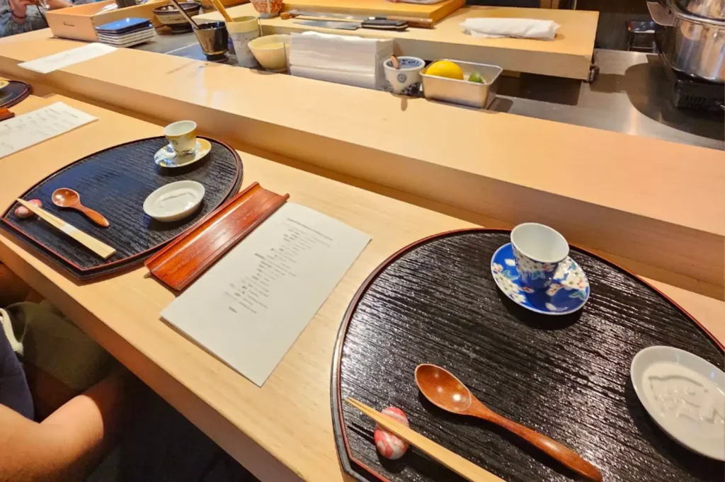 A high-angle shot shows two place settings arranged on a light wood sushi counter for an omakase meal. Each spot features a large, dark wood-textured tray holding a wooden spoon, a pair of chopsticks on a small patterned rest, a white dipping dish, and an ornate blue-and-white floral teacup on a matching saucer. A paper menu rests between the settings, while the raised chef's prep counter in the background holds containers of ingredients, sliced lemons, and kitchen linens.