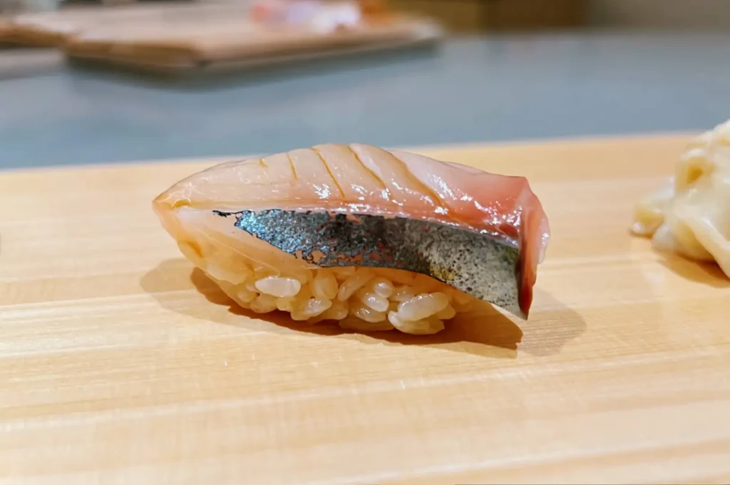 A piece of cured mackerel (shime saba) nigiri sushi featuring a shiny, patterned silver skin and scored top, served over seasoned sushi rice on a clean wooden dining counter next to a cluster of gari (pickled ginger).
