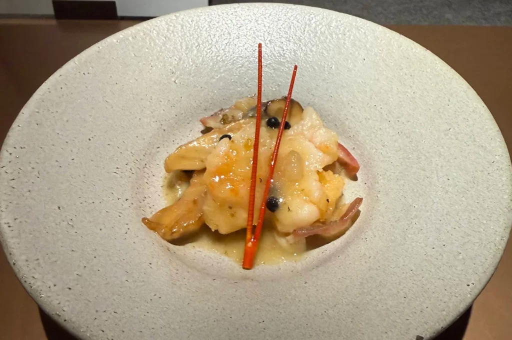 An elegant seafood dish presented in the center of a wide, textured light-grey ceramic bowl. The dish consists of tender, succulent pieces of cooked white fish or shellfish coated in a glossy, translucent sauce, tossed with slices of shiitake mushrooms and dark berries. Two long, ultra-thin, bright orange-red pine needles are crossed over the top as an artistic garnish, contrasting beautifully with the neutral tones of the bowl.