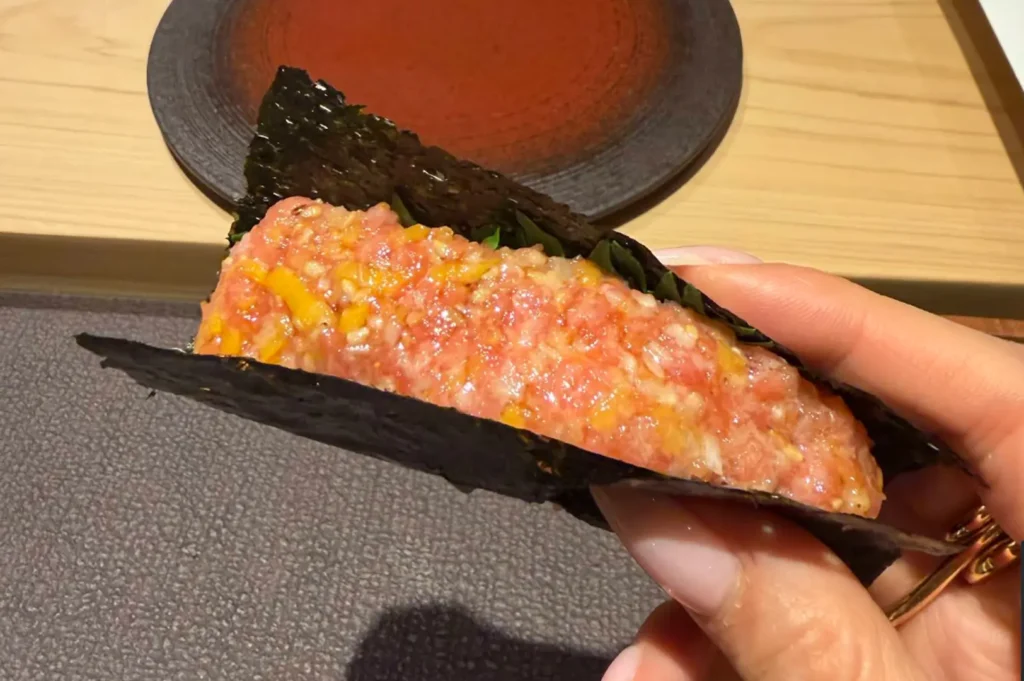 In this image, a hand holds a piece of sushi wrapped in a crisp sheet of nori, filled with a mixture of finely chopped raw fish and bright yellow ingredients, set against a blurred background of a wooden sushi counter and a dark, textured plate.