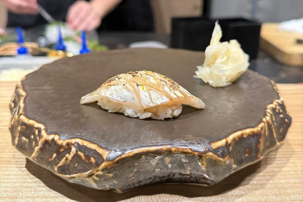 A close-up shot features a single piece of delicately seared nigiri sushi presented on a large, rustic ceramic plate with a textured, dark brown glazed surface and uneven edges. The sushi consists of a neat bundle of white rice topped with a slice of white fish that shows fine sear marks and a light glaze, garnished with tiny flecks of green zest. In the upper right background, a small, loose pile of pale pickled ginger sits as an accompaniment, while the distant background shows the blurred hands of a chef working at the sushi bar.