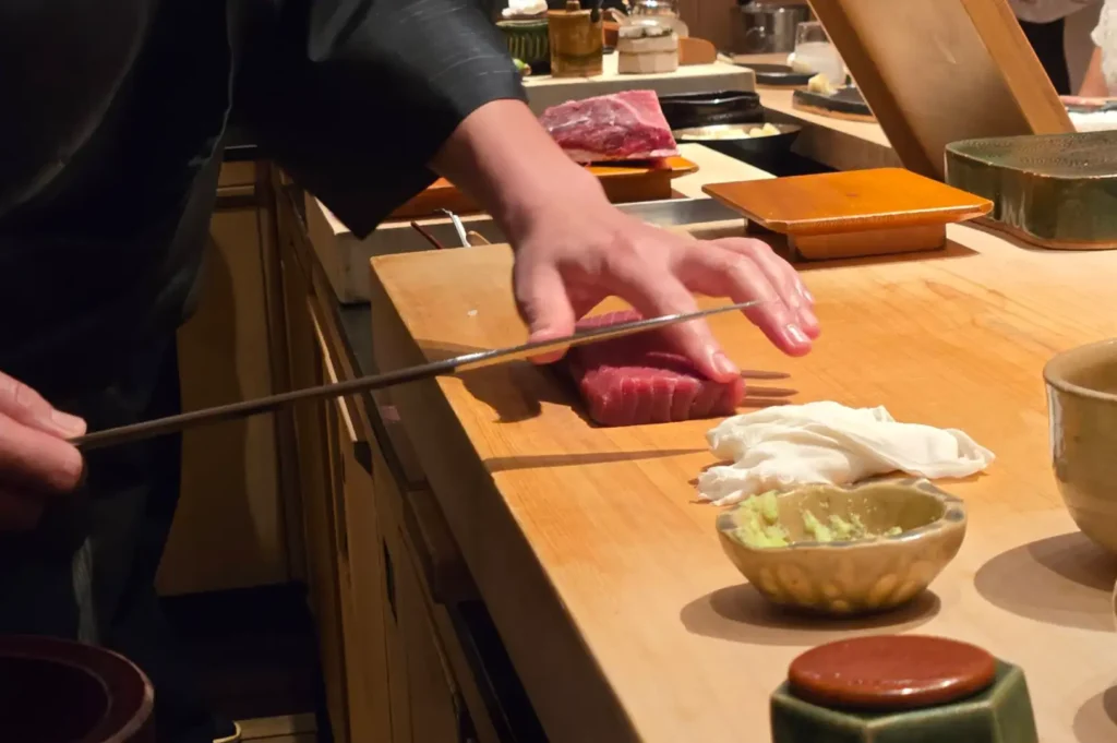 A chef dressed in dark clothing carefully sliced a thick piece of fresh red tuna on a wooden cutting board using a long, slender Japanese sushi knife, with small bowls of freshly grated wasabi nearby.