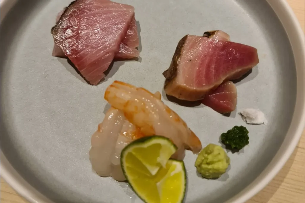 A light-grey plate holds an assortment of three types of sashimi, each distinct in color and texture, arranged alongside small, precise mounds of bright green wasabi and a dark green herb paste. A wedge of fresh lime and a small portion of coarse salt are placed near the fish, allowing for a customizable and refined tasting experience that highlights the natural flavors of the seafood.