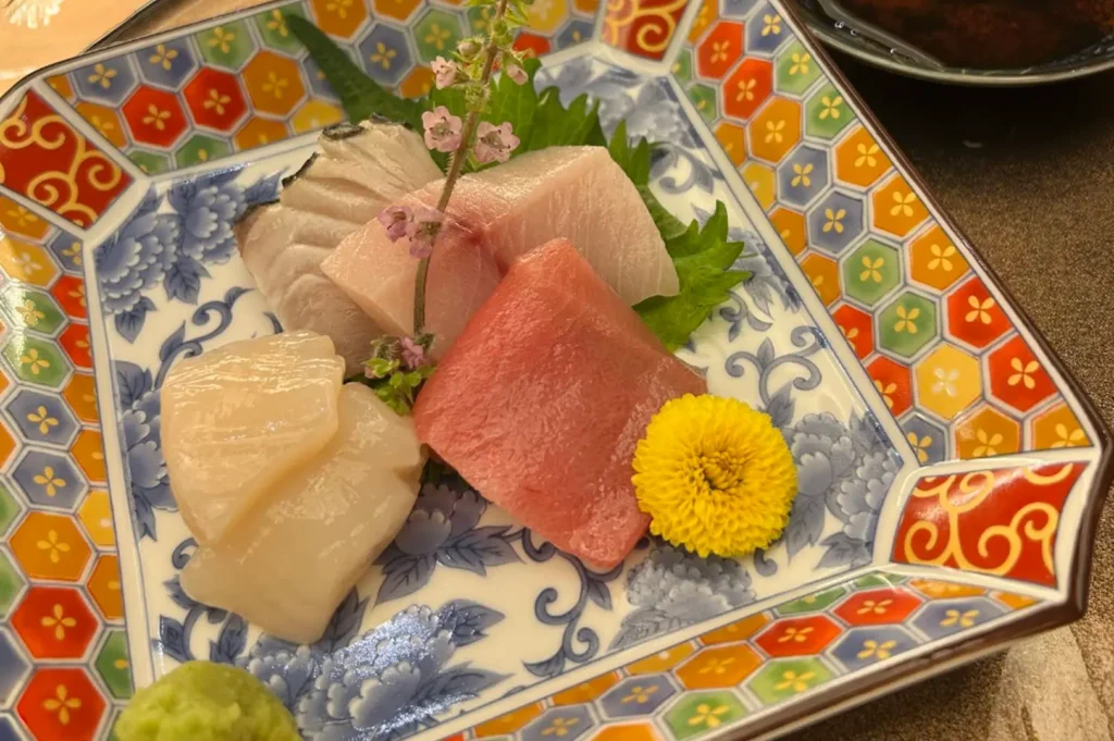 An assortment of premium sashimi, including scallops, tuna, and white fish, is presented on a decorative square plate featuring a colorful geometric honeycomb border and blue floral motifs, topped with a small, delicate sprig of flowers and a bright yellow chrysanthemum.