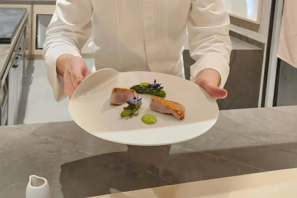 A sushi chef dressed in a pristine white chef jacket extends a wide, asymmetrical white ceramic plate directly toward the camera. On the minimalist plate are two thick cuts of cooked fish with golden, crispy seared skin, presented alongside charred asparagus spears, bright green puree dots, delicate microgreens, and small purple edible blossoms. The background shows a clean, modern omakase dining environment with light wood counters and warm ambient lighting.