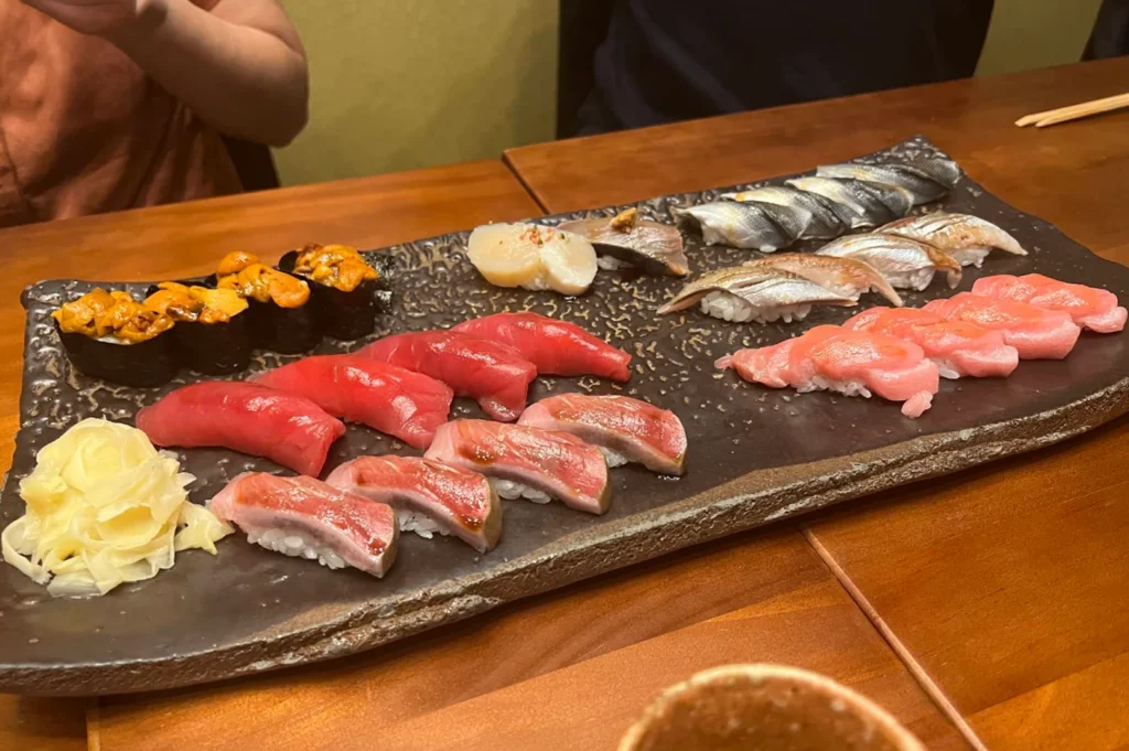 A wide, high-angle shot showing a large, rectangular black serving platter filled with an assortment of high-end nigiri sushi and a side of pickled ginger. The platter features varied pieces of seafood, including sea urchin (uni), bright red tuna, seared fish, and silver-skinned fish, artfully arranged on a wooden table in an intimate, softly lit restaurant setting.