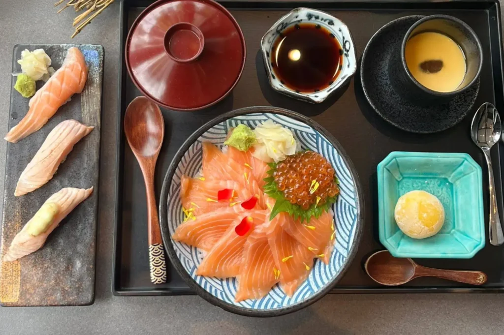 A black tray is meticulously arranged with a bowl of salmon sashimi over rice garnished with salmon roe, a side of salmon nigiri, a small cup of savory egg custard, a dish of soy sauce, and a round mochi dessert.