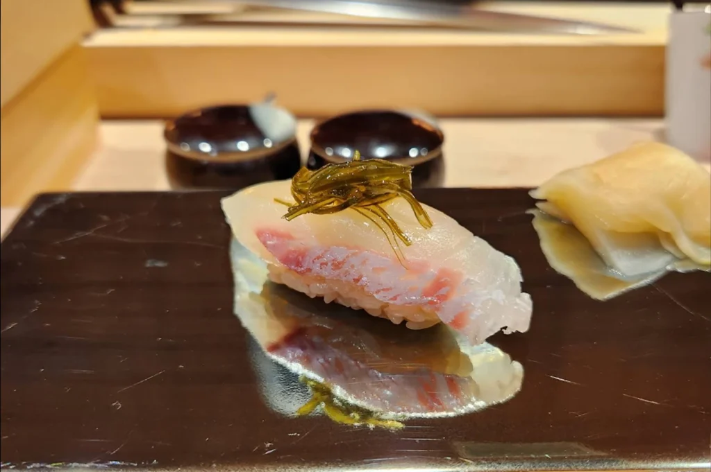 A close-up shot presents a piece of madai (sea bream) nigiri sushi resting on a sleek, reflective black lacquered platter. The translucent white fish showcases its distinct reddish-pink flesh underneath, pressed firmly onto a neat bed of seasoned sushi rice. A delicate bundle of dark green marinated kelp strings is placed elegantly on top of the fish as a garnish. To the right, a small portion of pale, folded pickled ginger sits as an accompaniment, while the glossy black platter mirrors the sushi and the dimly lit wooden counter elements in the soft-focus background.