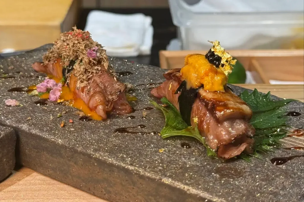 Two gourmet wagyu beef nigiri pieces are served on a dark stone slab, with one topped with shaved truffles and edible flowers, while the other is garnished with fresh sea urchin (uni), caviar, and edible gold leaf.