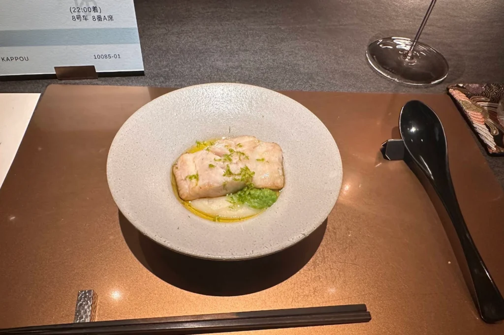 A minimalist and clean presentation of a cooked fish course served on a bronze placemat. A perfectly cooked, rectangular fillet of white fish rests on a smooth bed of white purée, garnished with a drizzle of yellow olive oil, a splash of vibrant green herb oil, and finely grated green citrus zest. A glossy black spoon on a dark rest and a pair of dark wooden chopsticks flank the wide, shallow grey bowl, with a corner of a custom paper ticket visible in the upper left.