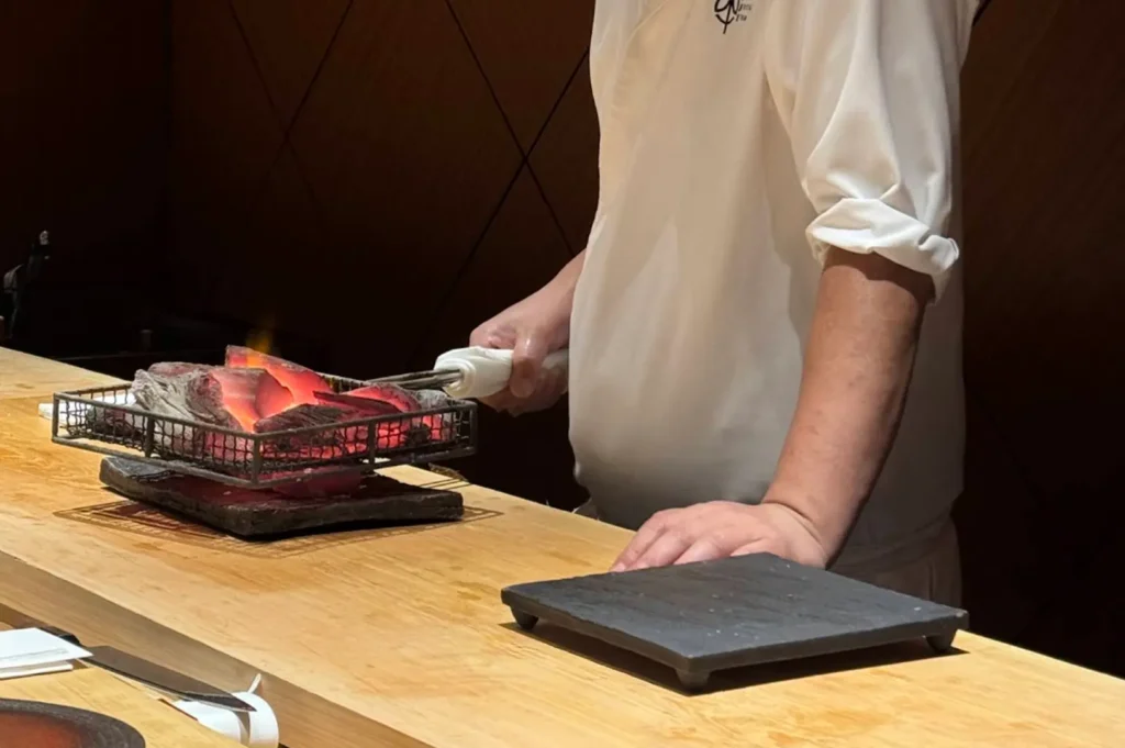 A sushi chef dressed in a white uniform stands behind a light-colored wooden counter, using tongs to manipulate a metal basket filled with glowing, hot charcoal for grilling, while a small black stone plate sits empty on the counter in the foreground.