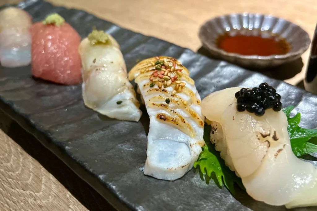 A close-up, angled view displays a selection of five distinct pieces of nigiri sushi neatly arranged in a row on a long, dark grey slate platter with a textured surface. Moving from right to left, the front piece features translucent white seafood topped with a cluster of shiny black caviar, followed by a piece of seared white fish drizzled with sauce and sprinkled with sesame seeds, a pale translucent fish topped with a dab of green wasabi, a piece of pink tuna with wasabi, and a partially visible pale piece at the end. In the soft-focus background, a small textured ceramic dish filled with dark soy sauce sits on the light wooden table.