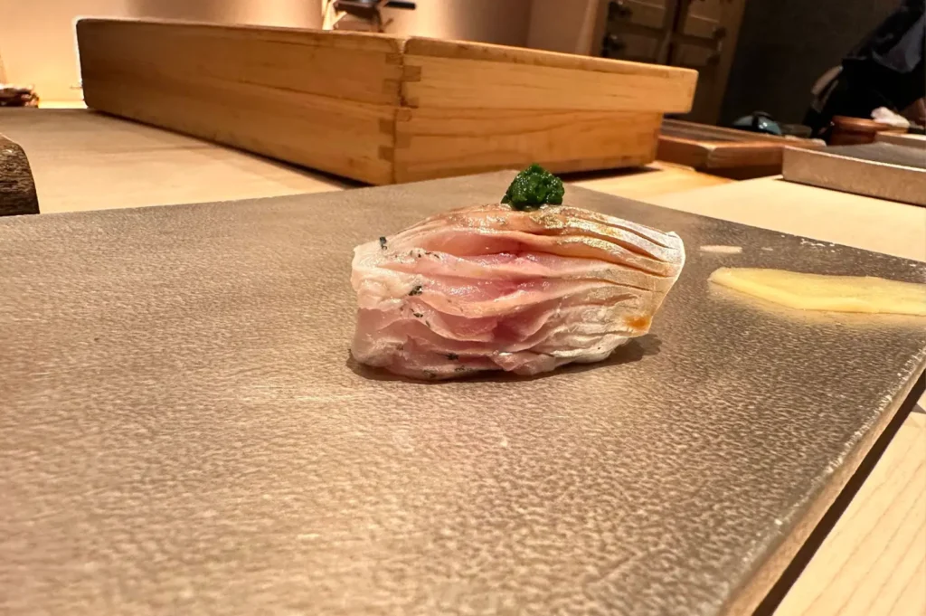 A piece of silver-skinned fish nigiri, scored with precision, rests on a textured stone serving board alongside a small piece of pickled ginger. A tiny, vibrant green garnish sits atop the fish, demonstrating the chef's attention to detail and commitment to elevating even the most traditional sushi with subtle, flavorful enhancements.