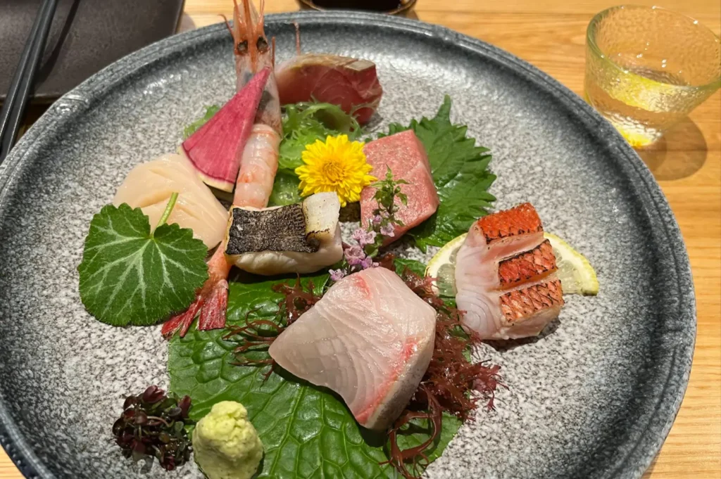 A gray stone-textured plate serves as the backdrop for a beautifully plated sashimi selection, featuring fresh shrimp, scallops, tuna, and seared fish, all garnished with shiso leaves, bright edible flowers, and a small mound of freshly grated wasabi.