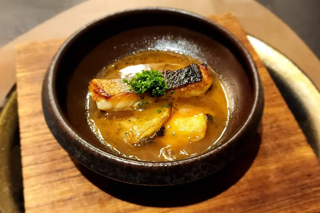 A detailed view of a hot omakase dish presented in a small, rustic dark brown ceramic bowl set atop a square wooden block. Inside the bowl, a piece of cooked fish with dark, flame-charred skin sits partially submerged in a rich, golden broth alongside tender slices of braised abalone or mushroom. The fish is finely garnished with a neat mound of chopped fresh chives, catching the warm overhead lighting.