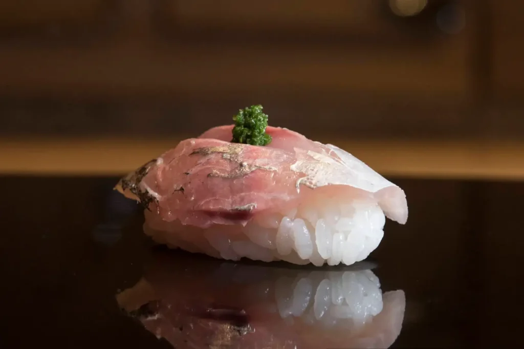 A single piece of delicate, translucent fish nigiri garnished with a small dab of green herb paste sits on a reflective black counter.