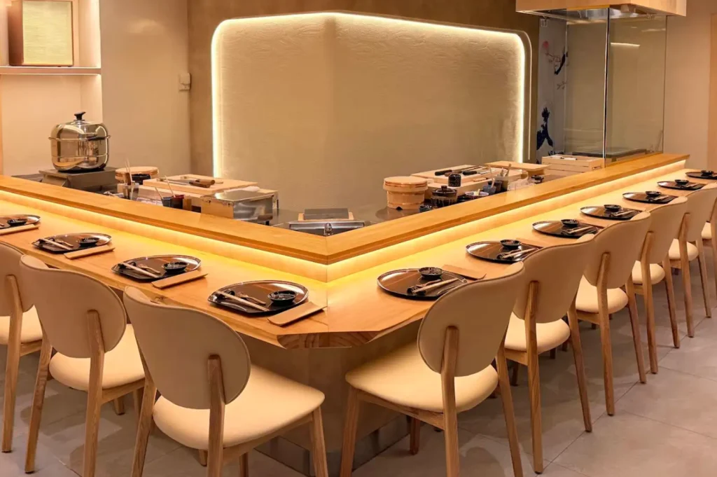 A sleek, modern Japanese dining space featuring a long, L-shaped light wood chef's counter warmly illuminated by built-in LED strip lighting, surrounded by minimalist beige upholstered chairs set with individual dark place settings, chopsticks, and soy sauce dishes.