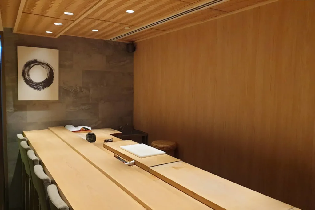 This image displays a minimalist, elegant sushi bar interior with a long, light-wood counter featuring several chairs, soft recessed lighting in the ceiling, a large piece of circular wall art, and a clean, understated aesthetic throughout the room.