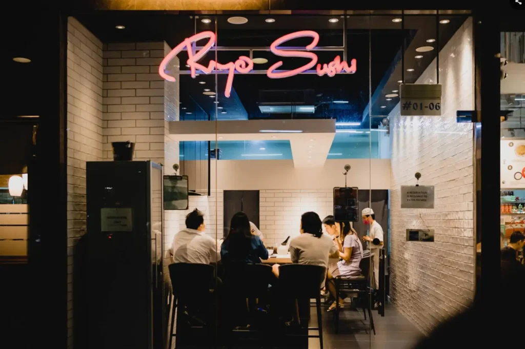A view from outside through glass panels shows the front of "Ryo Sushi," highlighted by a bright pink neon sign overhead and diners seated along a sleek sushi counter inside white brick walls.