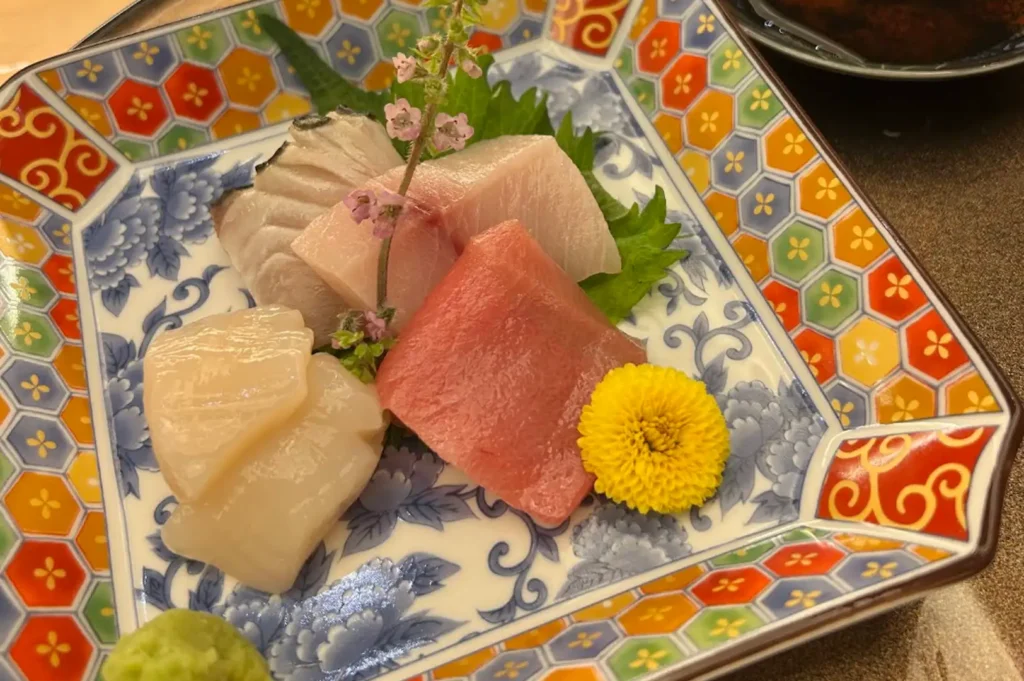 A top-angle view of an assortment of fresh sashimi—including thick cuts of raw tuna, scallop, yellowtail, and mackerel—served on a square ceramic plate with blue floral patterns and a colorful geometric border, garnished with a bright yellow chrysanthemum, green shiso leaf, delicate pink blossoms, and a dab of wasabi in the corner.