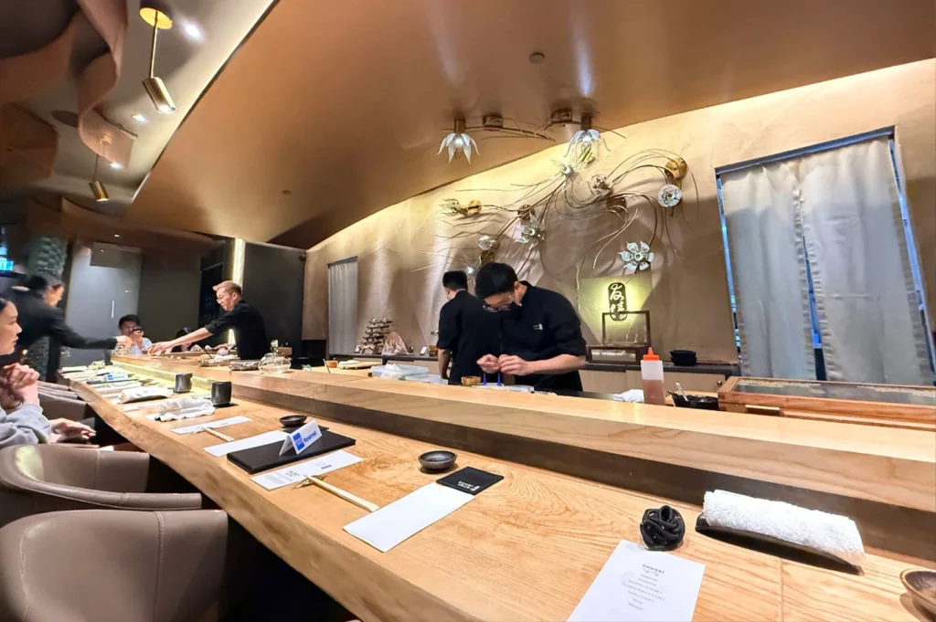 In a high-end restaurant with a gold-toned, curved ceiling and intricate floral wall art, chefs in black uniforms work behind a long, polished light-wood counter where guests are seated for an omakase experience.