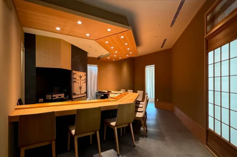 A wide interior view of an elegant, minimalist omakase dining room featuring a polished L-shaped light-wood sushi counter lined with modern wooden chairs with upholstered seats. Warm recessed lighting is embedded in an angled ceiling canopy above the counter, while earth-toned walls, dark flooring, a traditional shoji screen door on the right, and a traditional wooden icebox (nikkei) behind the prep area create a refined, serene atmosphere.