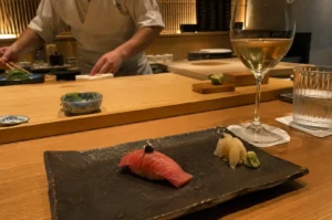 A diner's perspective at an upscale omakase counter, featuring a textured dark slate platter holding a piece of rich, pink fatty tuna (otoro) nigiri garnished with caviar, alongside pickled ginger and fresh wasabi. In the foreground sit a glass of white wine and a tumbler of water, while a sushi chef in a white uniform works attentively behind the smooth wooden counter in the warm, ambiently lit background.