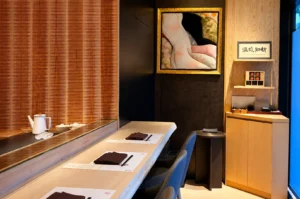 An interior view shows a minimalist Japanese omakase dining counter lined with smooth, light-colored wood tables, dark modern chairs, and neat place settings with folded brown napkins. In the background, a framed artwork, a Japanese calligraphy sign, and slatted wooden wall paneling complement the warm lighting and refined, intimate setting.