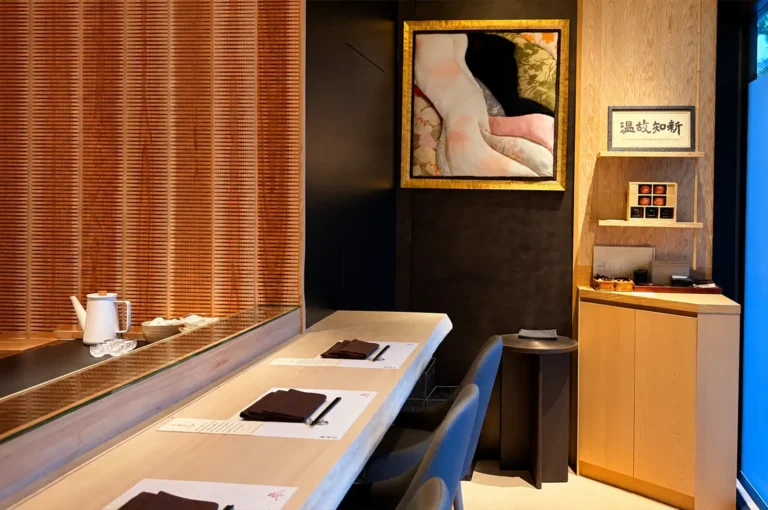 An interior view shows a minimalist Japanese omakase dining counter lined with smooth, light-colored wood tables, dark modern chairs, and neat place settings with folded brown napkins. In the background, a framed artwork, a Japanese calligraphy sign, and slatted wooden wall paneling complement the warm lighting and refined, intimate setting.