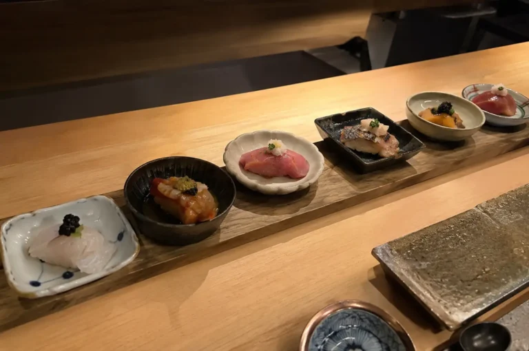 An eye-level shot captures a line of six artisanal omakase dishes arranged across a wooden platter on a light-toned wooden sushi counter. Each piece is individually served in a unique ceramic dish, featuring delicate toppings such as caviar, grated daikon, and fine herbs over various cuts of fish, including white fish, marinated shrimp, fatty tuna, seared mackerel, and uni. In the foreground, a textured rectangular stone plate rests on the counter alongside a small blue ceramic dish.