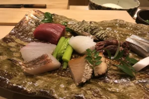 A close-up view of an omakase course presented on an earthy, textured, green-and-brown glazed ceramic platter resting on a light wooden counter. The arrangement features an assortment of fresh seafood and accompaniments, including thick slices of raw red tuna (maguro), pale white fish fillets—one with seared skin—bright green asparagus spears, translucent squid, tiny firefly squid (hotaru ika), tender sliced abalone next to its textured shell, and monkfish liver (ankimo) garnished with green leaves, with a chef's knife and ceramic bowls softly blurred in the background.