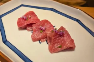 A close-up, top-down view of three delicate folds of richly marbled, pink otoro sashimi artfully arranged on a white hexagonal ceramic plate with a hand-painted blue border, each piece delicately garnished with tiny purple shiso flowers and micro-greens.